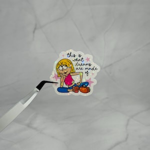 May include: A holographic sticker featuring a cartoon character with blonde hair, a pink top, and blue pants, sitting and resting their chin on a hand. The text reads "this is what dreams are made of" surrounded by small pink stars and sparkles.