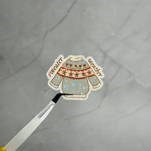 以下が含まれることがあります： 薄緑色のセーターの形をしたホログラムステッカーで、茶色の花とハートの模様が描かれています。セーターの両側には茶色の筆記体で「sweater weather」という文字が印刷されています。ステッカーには星が輝くようなエフェクトが施されています。