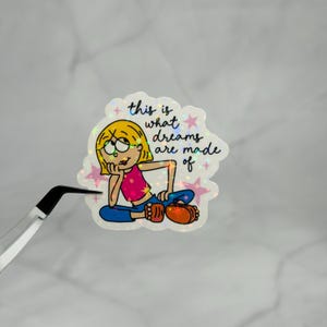 May include: A holographic sticker featuring a cartoon character with blonde hair, a pink top, and blue pants, sitting and resting their chin on a hand. The text reads, this is what dreams are made of, surrounded by small pink stars on a shimmering surface.