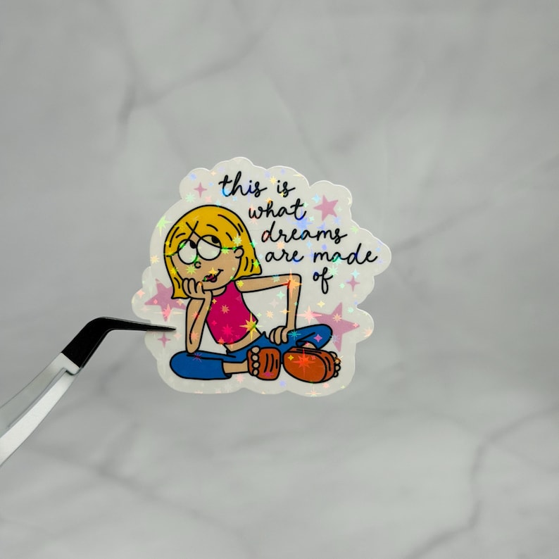 May include: A holographic sticker featuring a cartoon character with blonde hair, a pink top, and blue pants, sitting and resting their chin on a hand. The text reads, this is what dreams are made of, surrounded by small pink stars on a shimmering surface.