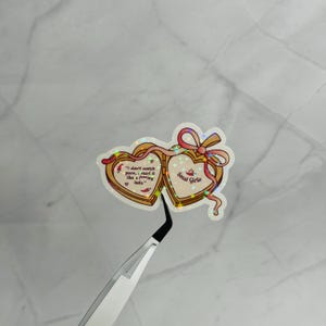 May include: A holographic heart-shaped locket sticker featuring a pink ribbon and the text: I don't watch porn, i read it like a f***ing lady. and Smut Girlie. The sticker has a gold-toned border and small red chili pepper illustrations.