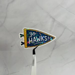 May include: A sticker in the shape of a pennant with the words "go HAWKS!" in white script. The pennant is navy blue with gold trim and a holographic finish. The sticker is held by a pair of tweezers.
