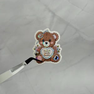 May include: A brown teddy bear sticker with a holographic finish featuring a light blue heart on its chest with the text Dearest Gentle Reader. The bear is adorned with a light blue bow, pink flowers, and a small bee illustration on its paw.