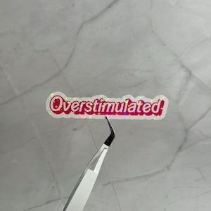 以下が含まれることがあります： ピンク色のバブルスタイルのフォントで「Overstimulated」という言葉が入ったホログラフィックステッカー。ステッカーは、大理石の背景に対して、銀色のピンセットで保持されています。ステッカーには白い縁があります。