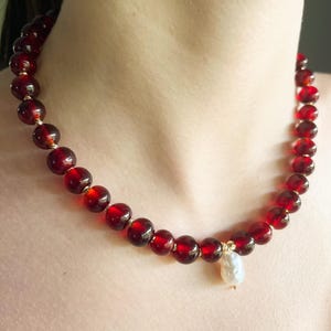 May include: A necklace featuring large, round, deep red beads strung together with gold-colored accents. A single, irregular-shaped white pearl pendant hangs from the center of the necklace. The necklace is displayed around a person's neck.