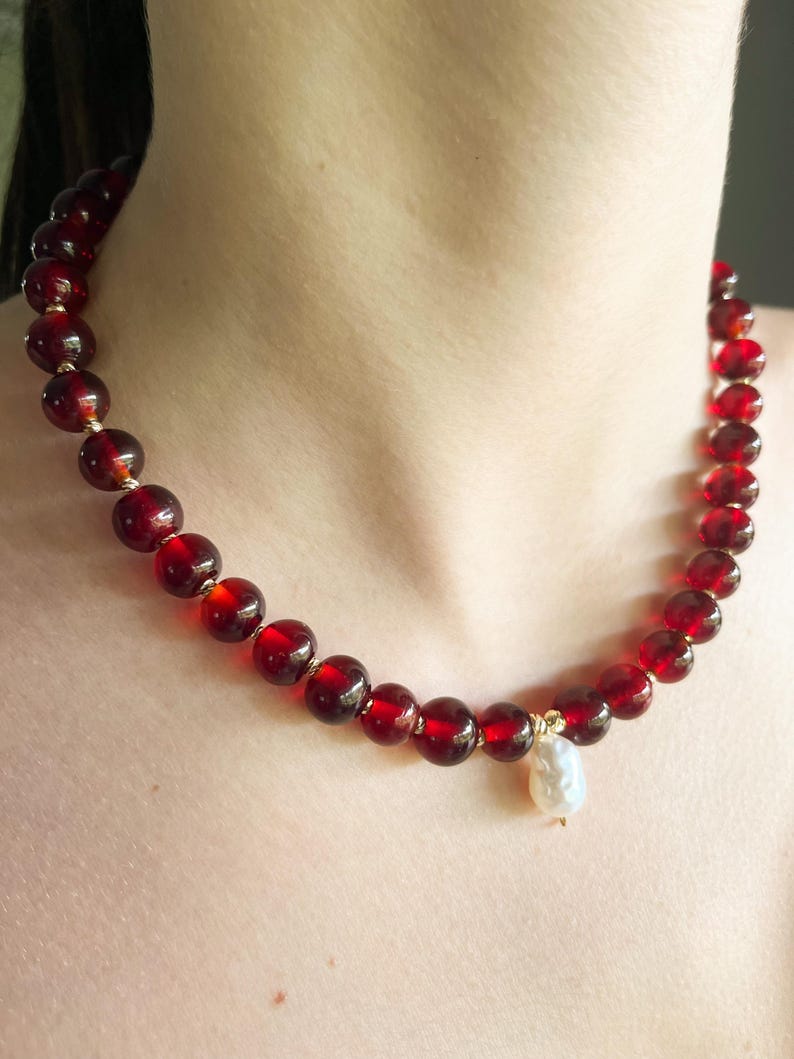 May include: A necklace featuring large, round, deep red beads strung together with gold-colored accents. A single, irregular-shaped white pearl pendant hangs from the center of the necklace. The necklace is displayed around a person's neck.