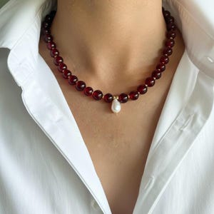May include: A beaded necklace featuring deep red, round beads. The necklace has small gold-colored spacers and a single, pear-shaped white pearl pendant. The necklace is worn against a white collared shirt.