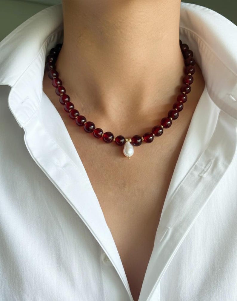 May include: A beaded necklace featuring deep red, round beads. The necklace has small gold-colored spacers and a single, pear-shaped white pearl pendant. The necklace is worn against a white collared shirt.
