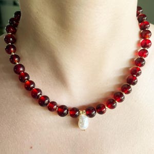 May include: A necklace featuring large, round, deep red beads strung together. The necklace has a gold clasp and a single, pear-shaped, white pearl pendant. The beads appear to be made of a translucent material.