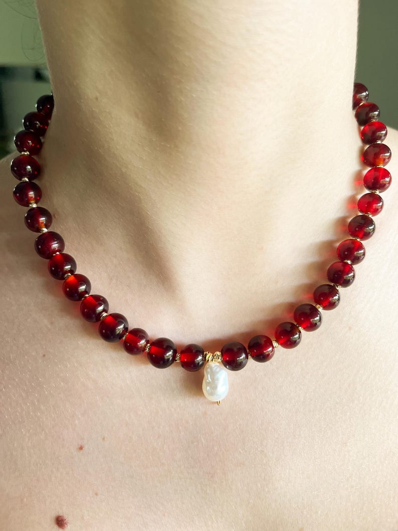 May include: A necklace featuring large, round, deep red beads strung together. The necklace has a gold clasp and a single, pear-shaped, white pearl pendant. The beads appear to be made of a translucent material.