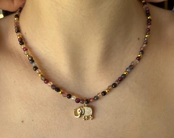 May include: A beaded necklace with a gold elephant pendant. The necklace features small, colorful beads in shades of red, purple, and gold. The elephant pendant is encrusted with small, clear stones.