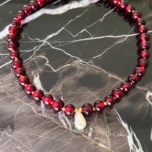 May include: A necklace featuring deep red, round beads strung together with gold-colored accents. A pear-shaped, off-white pendant hangs from the center. The necklace is displayed on a dark, marbled surface.