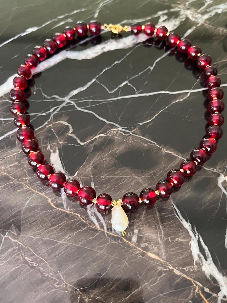 May include: A necklace featuring deep red, round beads strung together with gold-colored accents. A pear-shaped, off-white pendant hangs from the center. The necklace is displayed on a dark, marbled surface.