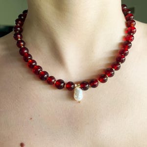 May include: A beaded necklace featuring large, round, deep red beads. The necklace has a gold-colored clasp and a single, white, teardrop-shaped pearl pendant. The necklace is worn around the neck.