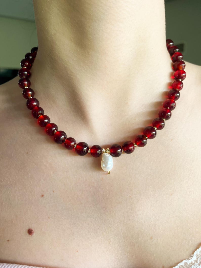May include: A beaded necklace featuring large, round, deep red beads. The necklace has a gold-colored clasp and a single, white, teardrop-shaped pearl pendant. The necklace is worn around the neck.