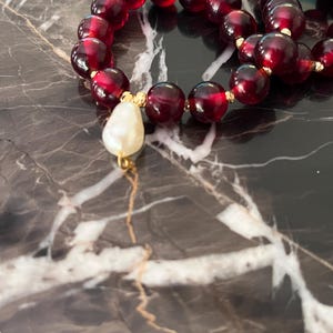 May include: A close-up of a beaded necklace featuring large, round, dark red beads. The necklace has gold-colored accents and a single, teardrop-shaped, off-white pearl pendant. The necklace is displayed on a dark, marbled surface.