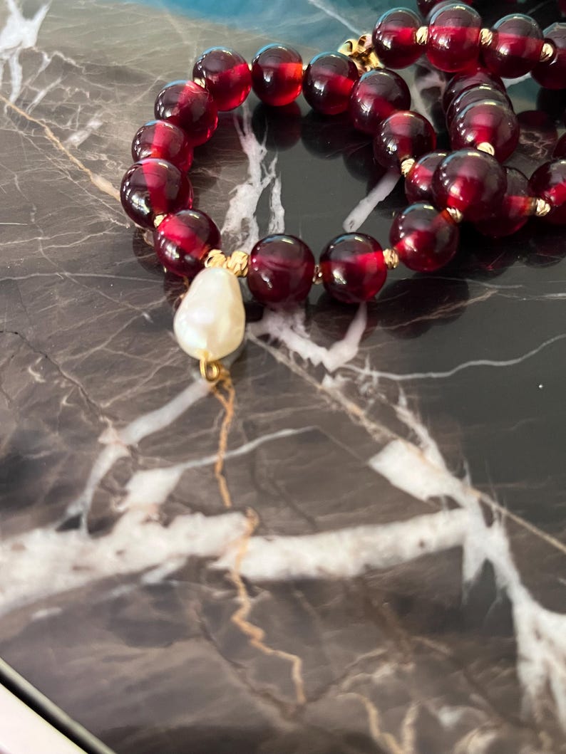 May include: A close-up of a beaded necklace featuring large, round, dark red beads. The necklace has gold-colored accents and a single, teardrop-shaped, off-white pearl pendant. The necklace is displayed on a dark, marbled surface.