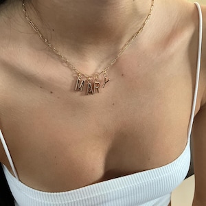 Könnte beinhalten: Eine goldfarbene Halskette mit dem Namen "MARY", der in einzelnen Buchstabenanhängern geschrieben ist. Die Halskette hat eine zarte Kette und die Buchstaben sind in einer einfachen Blockschrift gehalten. Die Halskette wird von einer Person getragen.