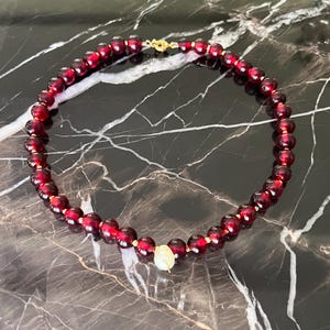 May include: A beaded necklace featuring deep red, round beads with gold-colored accents. The necklace has a gold clasp and a single, off-white, pearlescent bead as a focal point. The necklace is displayed on a dark, marble-like surface.