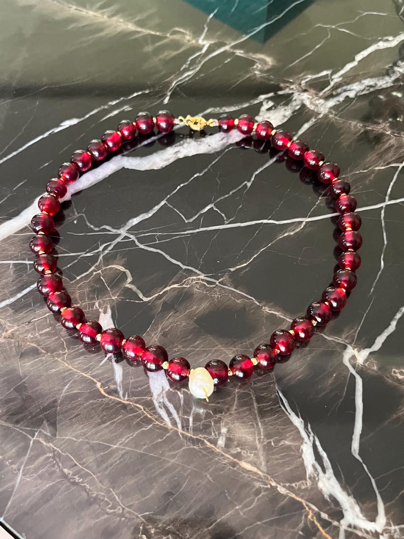 May include: A beaded necklace featuring deep red, round beads with gold-colored accents. The necklace has a gold clasp and a single, off-white, pearlescent bead as a focal point. The necklace is displayed on a dark, marble-like surface.