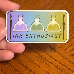May include: A sticker with a rainbow gradient background featuring three ink bottles in purple, green, and yellow. The bottles have a cork top and are filled with ink. The sticker also includes a quill pen and a paintbrush. The text "INK ENTHUSIAST" is printed below the bottles.