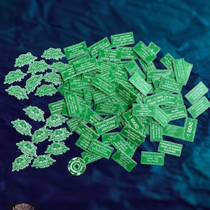 May include: A collection of green game tokens and markers on a dark blue surface. The tokens have various shapes and text, including words like "Polluted," "Diseased," and game-related terms. A few circular tokens are also present.