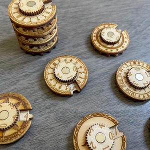 Könnte beinhalten: Ein Satz von sieben runden Holzteilen mit einem drehbaren Zifferblatt in der Mitte jedes Teils. Die Zifferblätter haben Zahlen und Symbole darauf. Die Teile sind auf einer hellbraunen Holzoberfläche angeordnet.