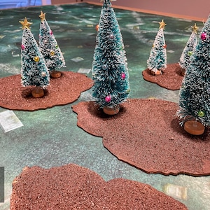 May include: Miniature Christmas trees with gold star toppers and colourful ornaments are arranged on brown, textured bases. The trees are set on a green and blue patterned surface, creating a festive holiday scene.