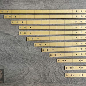 May include: A set of 12 gold-colored metal ruler strips with black markings. The strips are arranged in descending order of length, with the longest strip at the top and the shortest strip at the bottom.