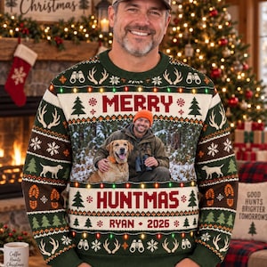 May include: A dark green long-sleeve knit jumper featuring a central photographic print of a person with a dog, surrounded by festive patterns of pine trees, antlers, binoculars, and Christmas lights, with the text MERRY HUNTMAS RYAN 2026.
