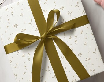 Puede incluir: Una caja de regalo envuelta en papel blanco con un patrón de pequeñas hojas marrones claras. Una cinta ancha de satén amarillo dorado está atada en un lazo en la parte superior de la caja, creando un acento decorativo.