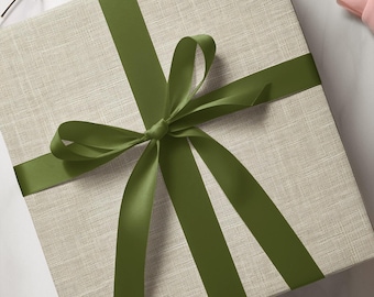 May include: A square gift box wrapped in light beige fabric with a textured linen-like finish. The box is tied with a smooth, dark olive green satin ribbon that forms a centred bow with four long, trailing ends extending across the top surface.