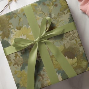 Puede incluir: Una caja de regalo cuadrada envuelta en papel con estampado floral con una cinta de raso verde claro atada en un lazo. El papel presenta un dise&ntilde;o de flores amarillas, verdes y marrones sobre un fondo verde claro.