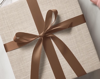 May include: A square gift box with a textured, light beige surface. A wide, brown satin ribbon is tied in a bow around the box. The bow is centered on the top of the box, creating a decorative and elegant presentation.