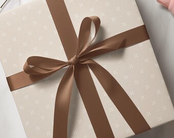 Puede incluir: Una caja de regalo cuadrada envuelta en papel beige claro con un patrón repetitivo de pequeñas cruces blancas. La caja está atada con una cinta de satén marrón oscuro lisa, dispuesta en un lazo central con cuatro extremos largos y caídos.