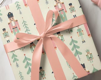 May include: A square gift box wrapped in light green paper featuring a repeating pattern of nutcracker figures in pink and green uniforms alongside stylised evergreen trees, tied with a wide, smooth, pale pink satin ribbon in a centred bow.