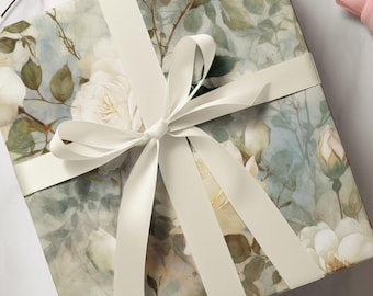 Peut inclure: Une boîte cadeau carrée enveloppée dans du papier à motif floral style aquarelle avec des roses blanches, des feuilles vertes et des touches bleu pâle. La boîte est nouée avec un ruban de satin blanc uni disposé en un nœud croisé classique.