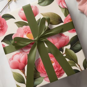 May include: A wrapped gift with floral wrapping paper featuring pink camellias and green leaves. The gift is tied with a dark green satin ribbon and bow, creating a visually appealing presentation.