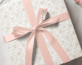 Peut inclure: Un coffret cadeau emballé dans du papier à motifs floraux, noué avec un ruban de satin rose clair. Le papier d'emballage présente un fond blanc et un motif de roses beiges.