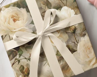 May include: A square gift box wrapped in paper featuring a detailed print of cream-colored roses, green leaves, and closed buds. The box is tied with a smooth, solid off-white satin ribbon arranged in a classic cross pattern with a centered bow.