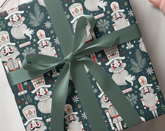 May include: A square gift box wrapped in dark teal paper featuring a repeating pattern of white nutcracker figures with red accents, pine branches, and small stars. The box is tied with a wide, smooth, sage green satin ribbon in a bow.