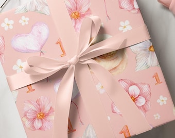 May include: A square gift box wrapped in light pink paper featuring a repeating pattern of watercolor-style pink flowers, heart-shaped balloons, and the number one in orange. The box is tied with a smooth, light pink satin ribbon bow.