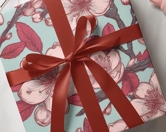 May include: A wrapped gift box featuring a floral pattern of pink blossoms and red leaves on a light blue background. A dark red satin ribbon is tied in a bow around the box, creating a decorative presentation.