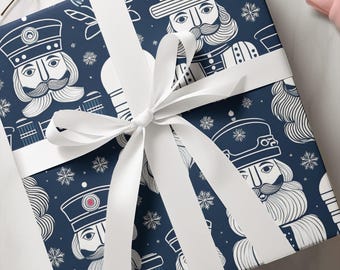 May include: A square gift box wrapped in dark blue paper featuring a repeating white line art pattern of nutcracker figures and snowflakes. The package is tied with a wide, smooth white satin ribbon arranged in a decorative bow on top.