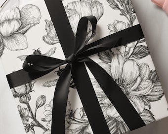 Puede incluir: Una caja de regalo cuadrada envuelta en papel blanco con una ilustración botánica detallada en tinta negra de flores y abejas. La caja está atada con una cinta de satén negra lisa dispuesta en un patrón de cruz con un lazo central.