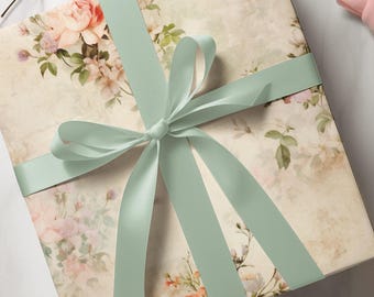 Peut inclure: Une boîte cadeau carrée enveloppée dans un papier couleur crème orné d'un motif floral délicat de roses roses et pêche avec des feuilles vertes. La boîte est nouée avec un large ruban de satin vert sauge disposé en un nœud centré.