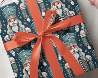 May include: A square gift box wrapped in dark teal paper featuring a repeating pattern of nutcracker figures with white beards and blue hats, accented by small white snowflakes. The box is tied with a wide, vibrant coral-coloured satin ribbon.