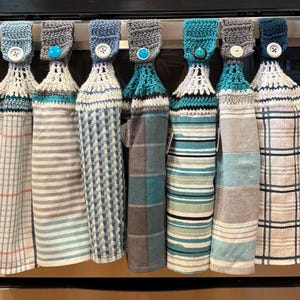 May include: Seven hanging kitchen towels with crocheted tops in shades of blue, gray, and white. Each towel features a button closure at the top. The patterns include stripes, plaids, and checks. One towel has a small white tag with the number 0847.