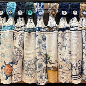 May include: Nine hanging kitchen towels with crocheted tops in various blue, white, and tan shades. Each towel features a button closure and a coastal-themed print including seashells, starfish, coral, a sea turtle, and a beach scene.