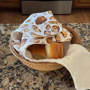 May include: A round woven wicker basket containing several golden-brown dinner rolls, covered by a cream-colored fabric cloth featuring a repeating pattern of various baked bread items including loaves, rolls, and croissants in shades of brown.